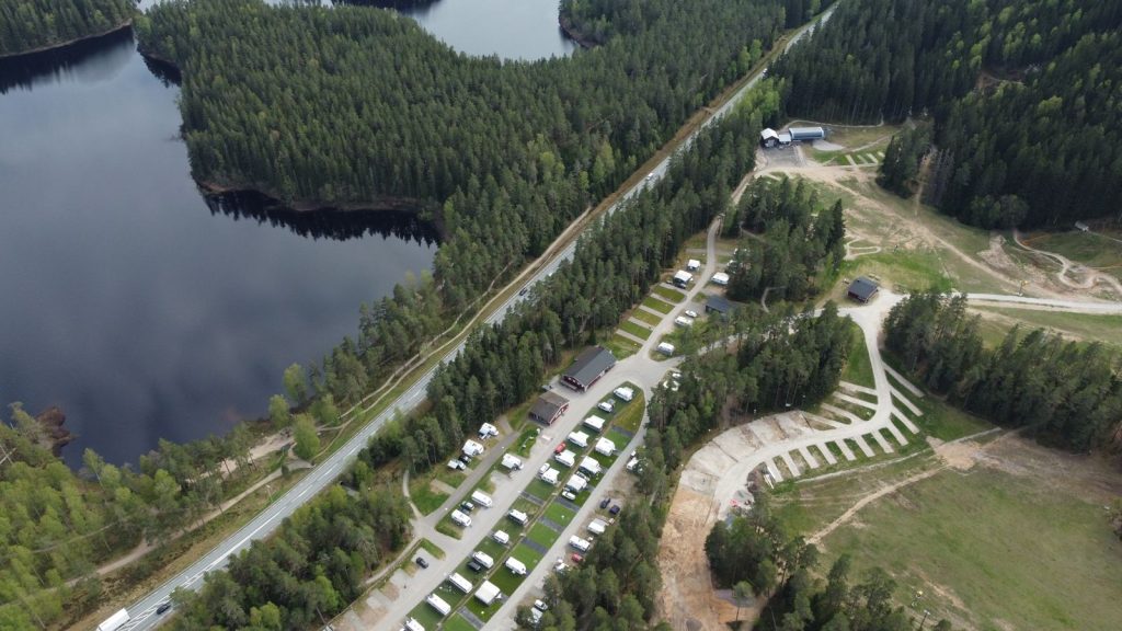 Luftaufnahme eines Campingplatzes an einem See mit Stellplätzen in Reihen zwischen Wald und Zufahrtswegen